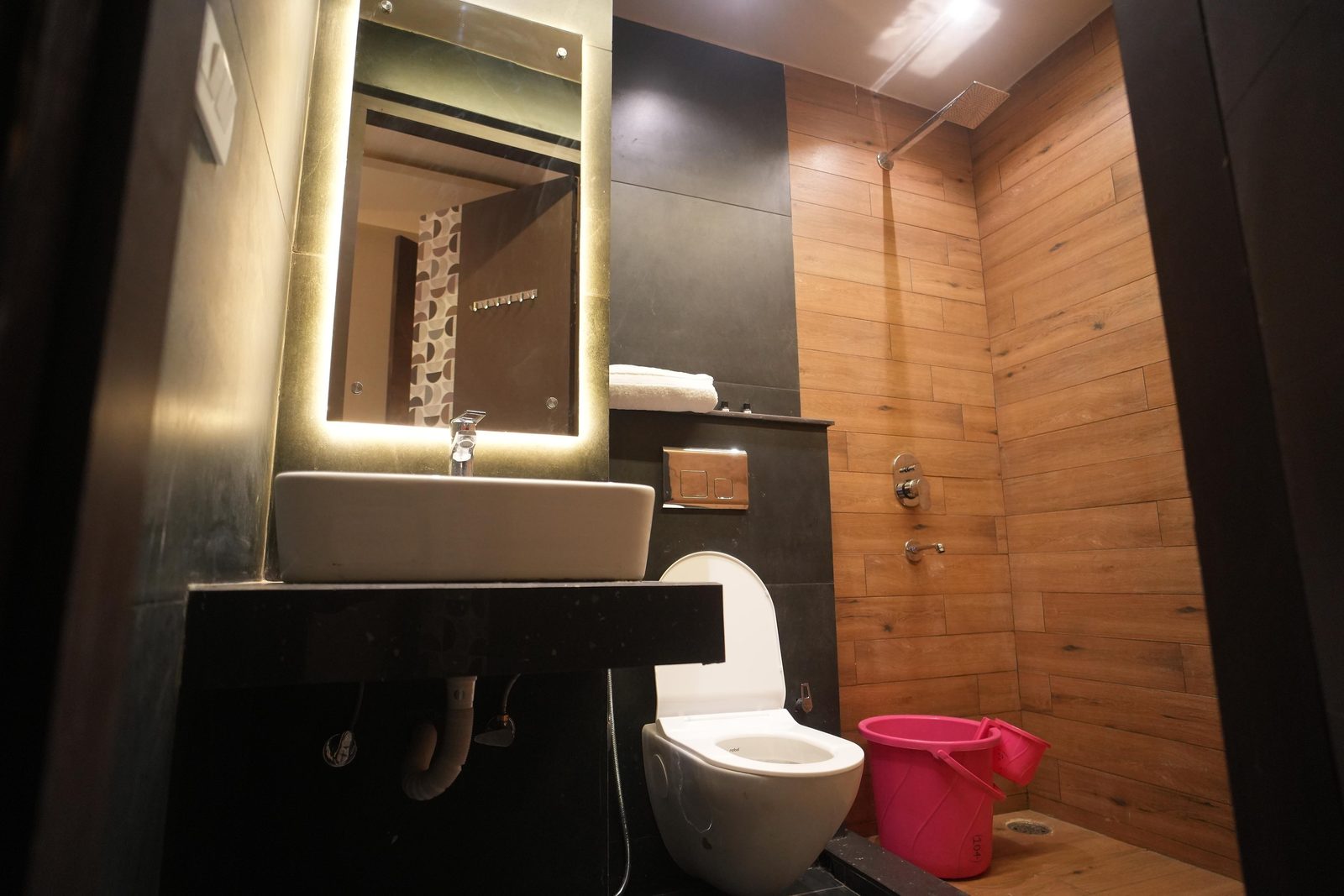 A dark-tiled bathroom with a backlit mirror over a vessel basin, a wall-hung toilet and a wood-panelled shower area.