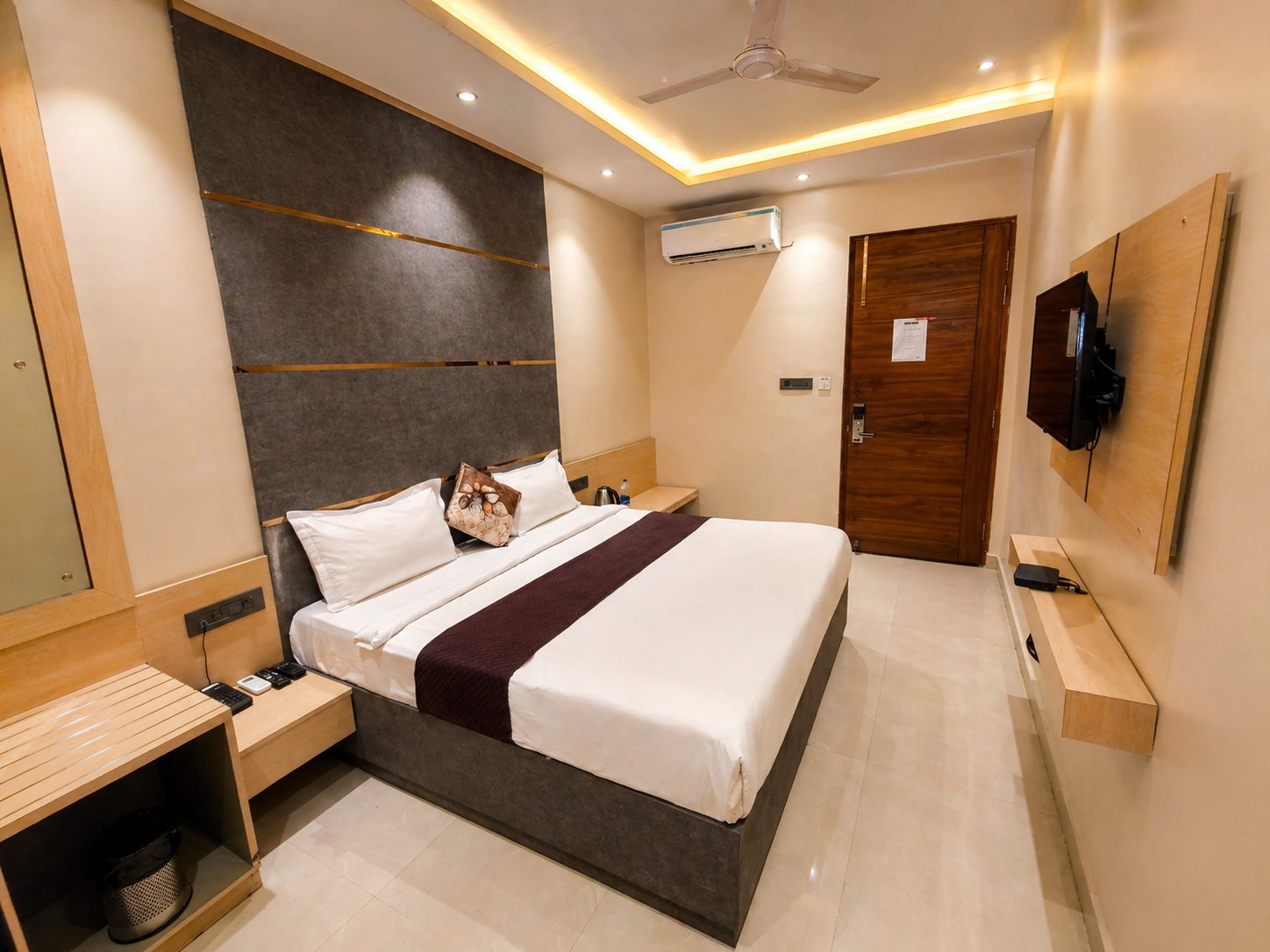 A guest room with a king bed against a grey felt wall inlaid with brass lines, a plum runner on white linen, a wall-mounted television and cove lighting.